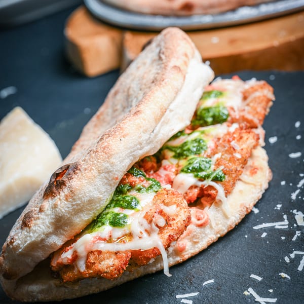 A crispy chicken sandwich with melted cheese and green pesto on a dark slate surface.