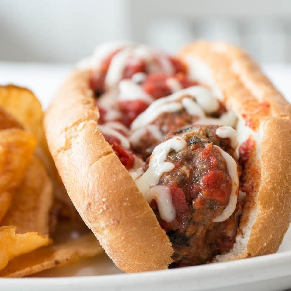 Meatball sub sandwich with melted cheese, marinara sauce, and a side of potato chips.