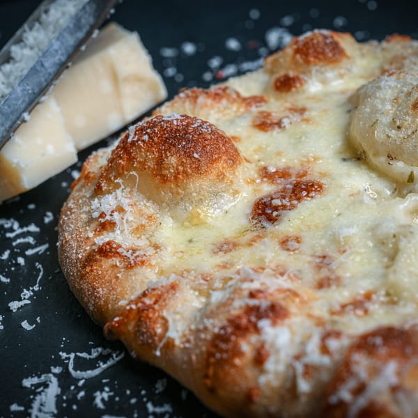 Close-up of a cheesy flatbread with a cheese grater and parmesan wedge in the background.