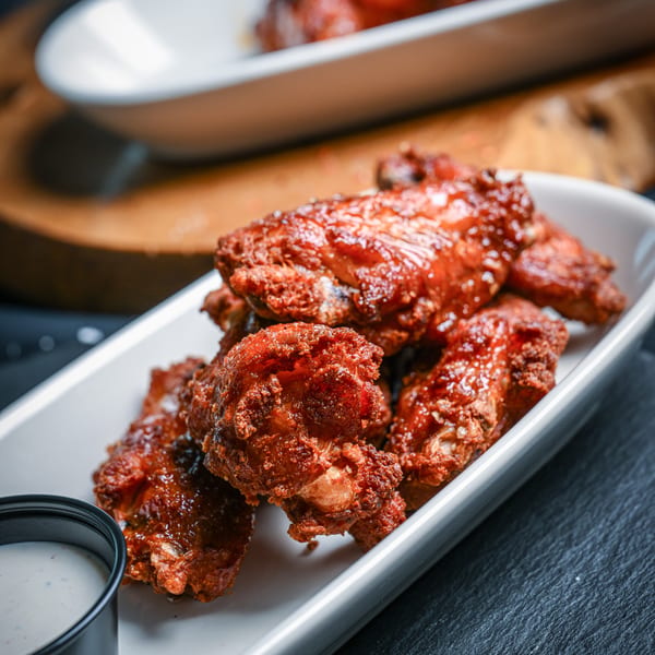 Crispy, saucy chicken wings in a white dish with a side of ranch dressing on a dark surface.